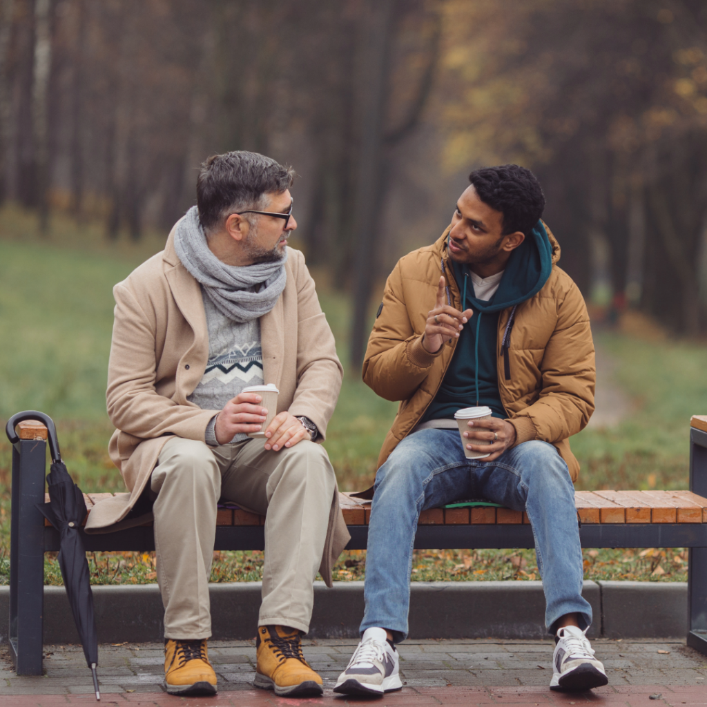 Two men talking over coffee on a park bench, starting an honest conversation after baby loss.